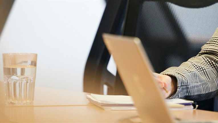 Laptop on desk zoomed in, person's hand writing in notebook