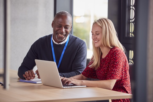 Two people sat a table in an office, smiling and looking at a laptop screen 